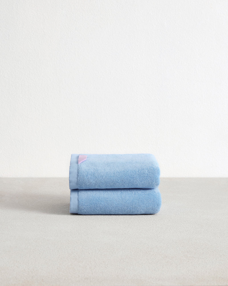 A set of two Sky 100% Cotton Terry Hand Towels by Bed Threads, in light blue, are neatly folded and stacked on a neutral surface with a white textured wall background.