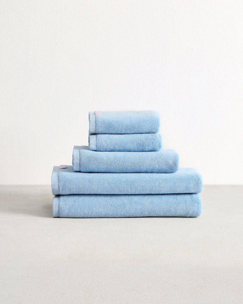 The Bed Threads Sky 100% Cotton Terry Bath Towel Bundle features five highly absorbent, light blue towels in assorted sizes, neatly stacked on a light surface against a plain white background.