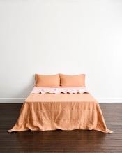 A neatly made bed with a peach-colored quilt cover in Terracotta & Rosewater, and two matching pillows in Terracotta, set against a plain white wall and dark wood floor. - view