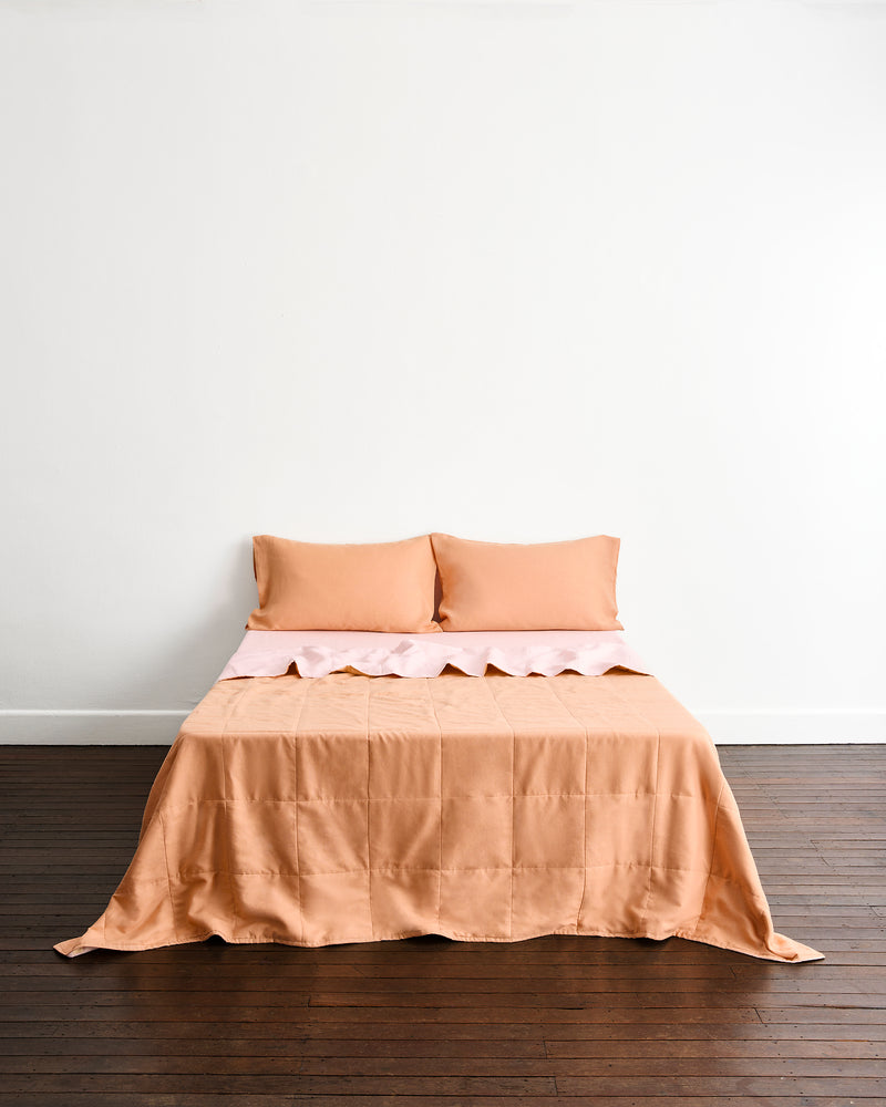A neatly made bed with a peach-colored quilt cover in Terracotta & Rosewater, and two matching pillows in Terracotta, set against a plain white wall and dark wood floor.