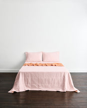 A neatly made bed with a light pink quilt cover in Terracotta & Rosewater and matching pillowcases in Rosewater, set against a plain white wall and dark wood floor. - view