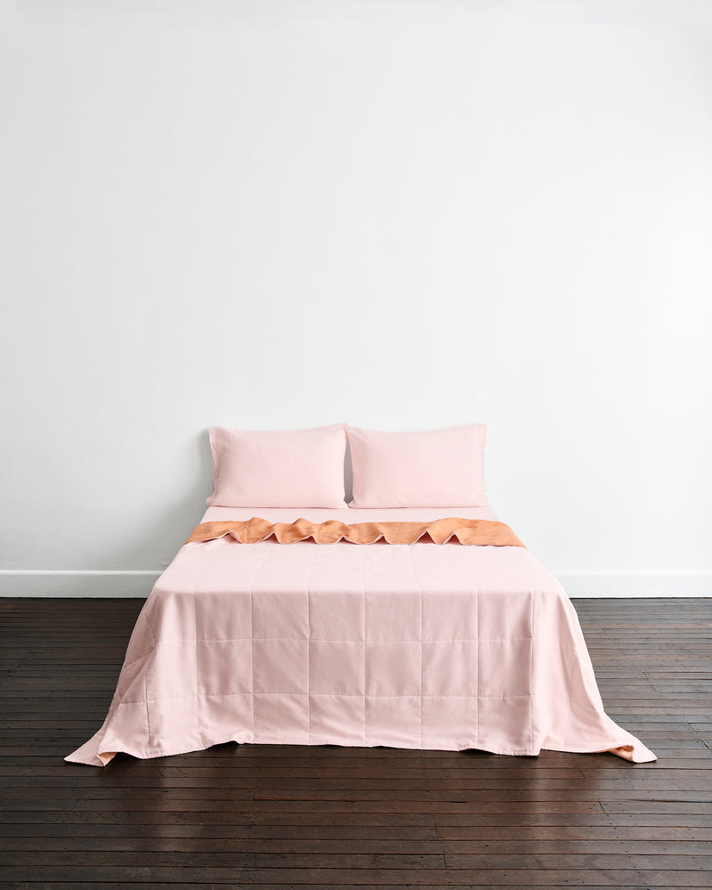 A neatly made bed with a light pink quilt cover in Terracotta & Rosewater and matching pillowcases in Rosewater, set against a plain white wall and dark wood floor.