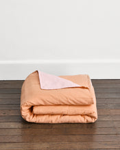 A folded peach-colored quilt cover in Terracotta & Rosewater with a light pink underside, displayed on a dark wood floor against a white wall. - view