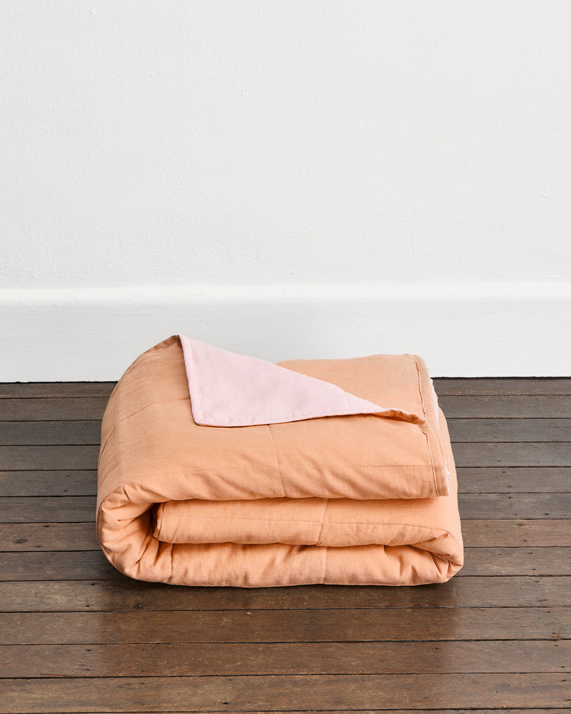 A folded peach-colored quilt cover in Terracotta & Rosewater with a light pink underside, displayed on a dark wood floor against a white wall.