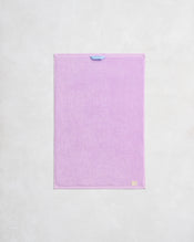The Violet 100% Cotton Terry Hand Towel by Bed Threads is a rectangular, pastel purple towel with a small blue loop, plush 650GSM texture, and a beige label in the corner, hanging against a light textured wall. - view
