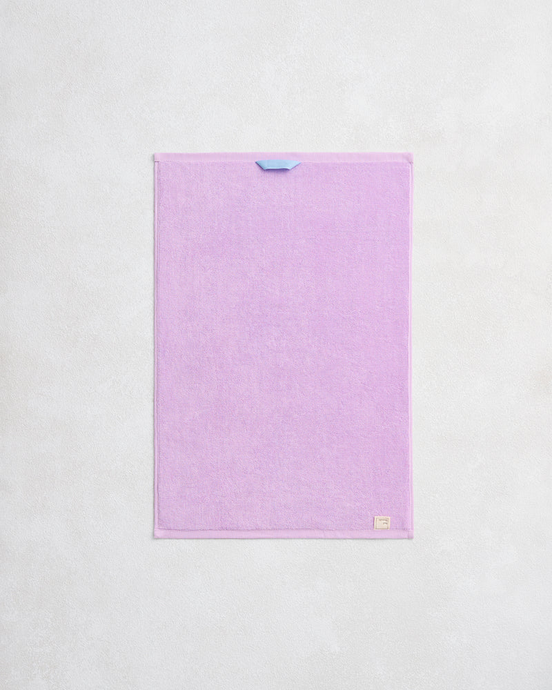 The Violet 100% Cotton Terry Hand Towel by Bed Threads is a rectangular, pastel purple towel with a small blue loop, plush 650GSM texture, and a beige label in the corner, hanging against a light textured wall.