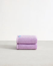 A pair of Violet 100% Cotton Terry Hand Towels by Bed Threads, featuring high absorbency and a small blue tag, is neatly folded on a light grey surface against a plain white background. - view
