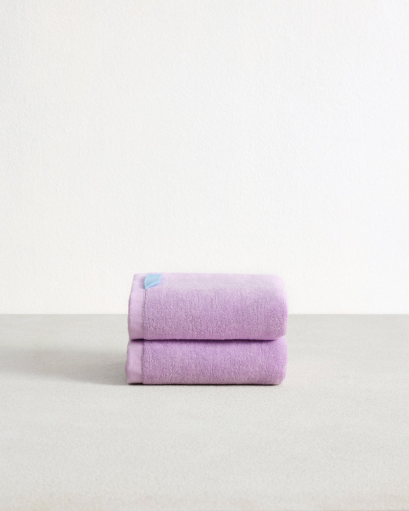 A pair of Violet 100% Cotton Terry Hand Towels by Bed Threads, featuring high absorbency and a small blue tag, is neatly folded on a light grey surface against a plain white background.