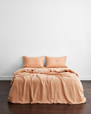 A neatly made bed features the Bed Threads Terracotta 100% French Flax Linen Bedding Set with two matching pillows, set against a plain white wall on a dark wooden floor.