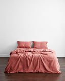 A neatly made bed features two pillows and a duvet in the Pink Clay 100% French Flax Linen Bedding Set by Bed Threads, set against a plain white wall and dark wooden floor.