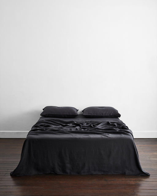 A neatly made bed features the Bed Threads Charcoal 100% French Flax Linen Flat Sheet and two black pillows, set against a plain white wall and a dark wooden floor.