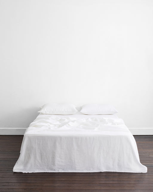 A double bed features the Bed Threads White 100% French Flax Linen Flat Sheet and two pillows, set against a plain white wall on a dark wooden floor in a minimalistic, uncluttered room.