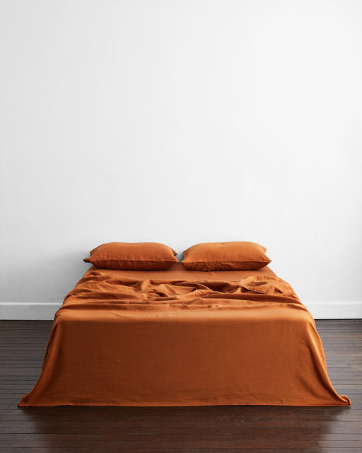 A neatly made bed features the Bed Threads Rust 100% French Flax Linen Flat Sheet with two matching pillows, set against a plain white wall on a dark wooden floor in a minimalistic room.