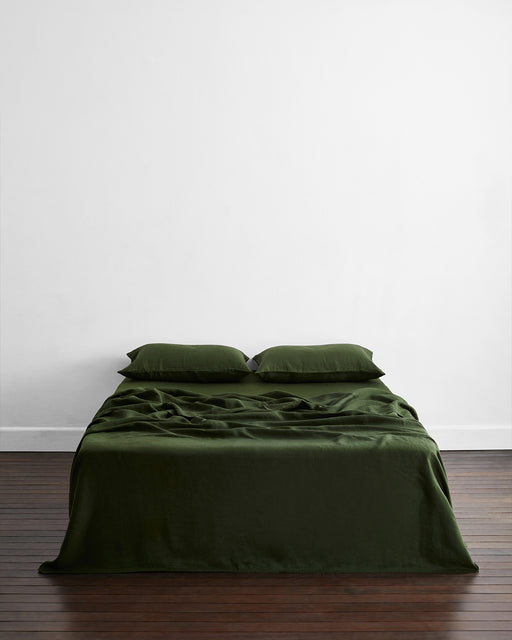 A minimalist room features a neatly made bed dressed with the Olive 100% French Flax Linen Flat Sheet by Bed Threads and two matching pillows, set on a wooden floor against a plain white wall.