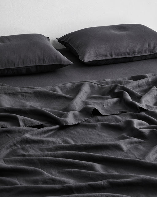 Two dark gray pillows and a Bed Threads Charcoal 100% French Flax Linen Flat Sheet rest on a bed against a plain white wall. The bedding has a smooth, matte texture, offering a soft and comfortable feel.