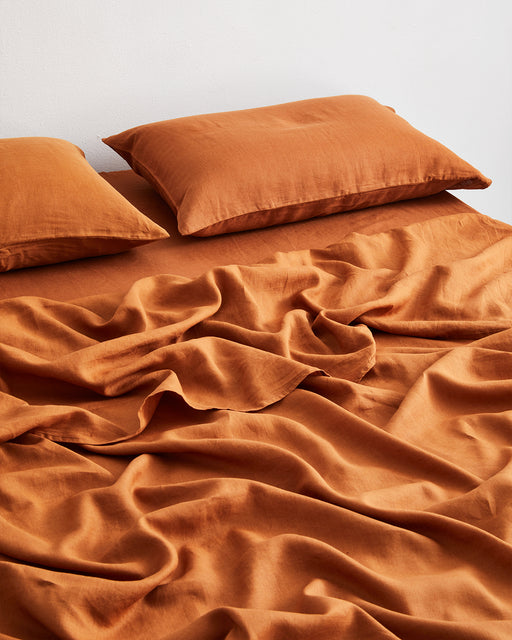 Two orange pillows and a Bed Threads Rust 100% French Flax Linen Flat Sheet, featuring a naturally wrinkled texture, are displayed on a bed against a plain white wall.
