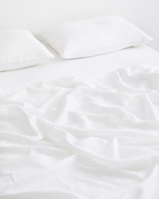 A neatly made bed features two white pillows and the Bed Threads White 100% French Flax Linen Flat Sheet, slightly wrinkled, set against a plain white background.