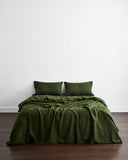 A minimalist room features a neatly made bed with two pillows and the Bed Threads Olive 100% French Flax Linen Bedding Set, topped with a green blanket, set against a white wall on a dark wooden floor.