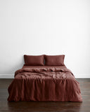 A minimalistic room features a neatly made bed with the Cacao 100% French Flax Linen Duvet Cover by Bed Threads and two pillows, set against a plain white wall on a dark wooden floor.